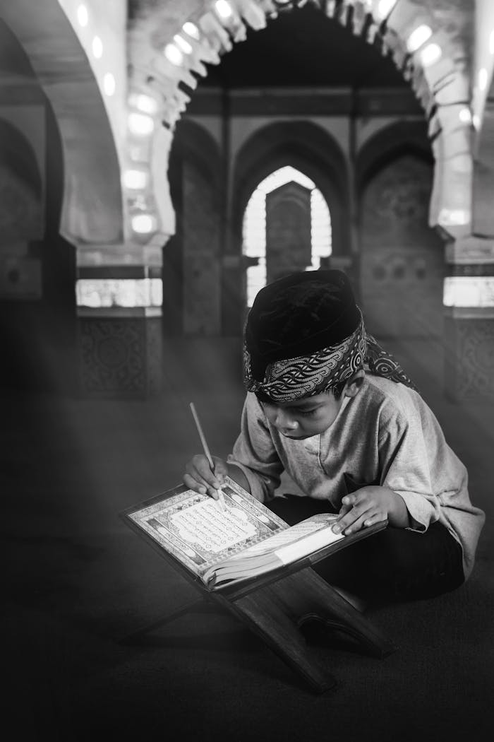 gallery-4 A young child deeply engaged in reading the Quran inside a mosque, captured in black and white.
